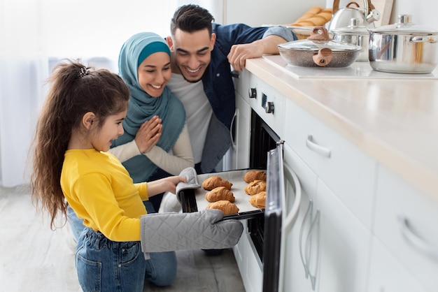 Glückliche muslimische dreiköpfige Familie, die zusammen in der Küche backt, süße kleine Tochter, die ein Tablett mit frisch gebackenen Croissants aus dem Ofen nimmt, fröhliche Eltern aus dem Nahen Osten und Kinder, die hausgemachtes Gebäck genießen