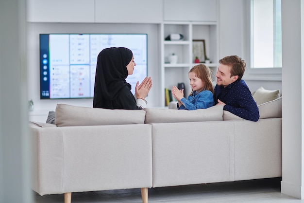 Glückliche muslimische Familie, die Zeit zusammen in einem modernen Zuhause verbringt. Eltern spielen während des Ramadan mit ihrer Tochter auf dem Sofa. Traditionelle modische arabische Kleidung.