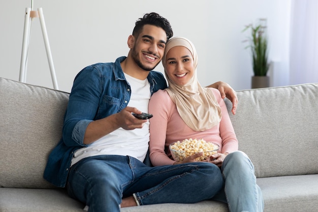 Glückliche muslimische zweiköpfige Familie, die zu Hause zusammen fernsehen, Popcorn umarmen und essen, Platz kopieren. Fröhlicher Mann und Frau aus dem Nahen Osten, die gemeinsam Spaß am Wochenende haben, Liebes- und Beziehungskonzept