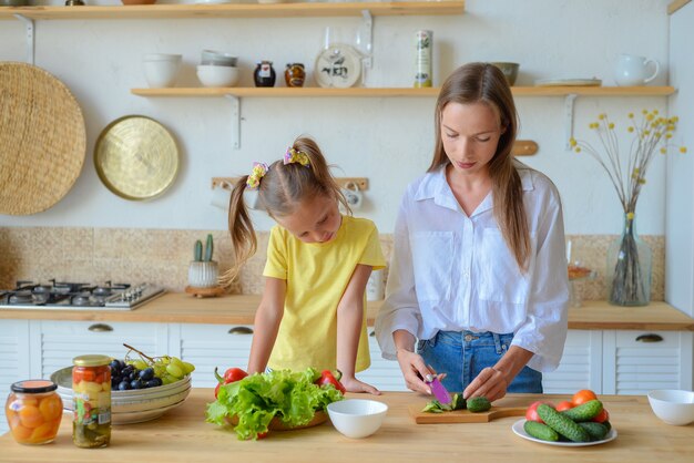 Glückliche Mutter bringt der kleinen Tochter bei, gesundes Essen zu kochen, Mutter und Mädchen, die lächelnd sprechen