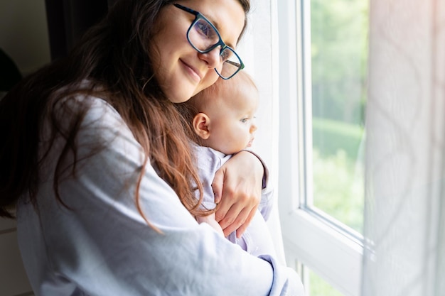 Glückliche Mutter, die ihre kleine Tochter hält und umarmt Mutter mit Baby, das in der Nähe des Fensters bleibt Glückliches Erziehungskonzept