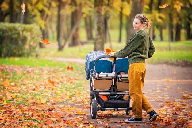 Glückliche Mutter, die im Herbstpark mit einem Kinderwagen für Zwillinge geht