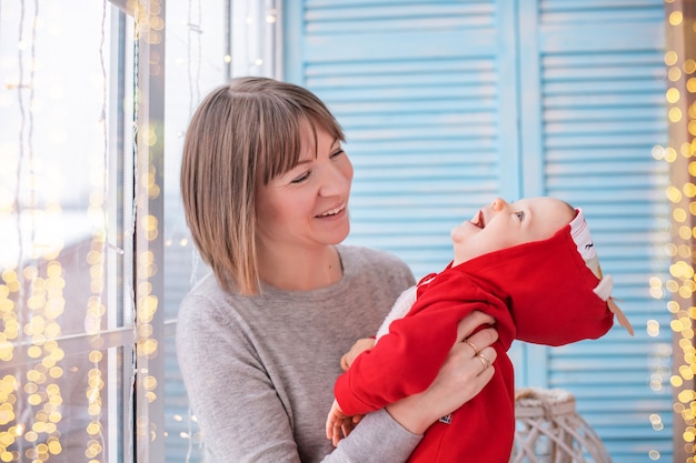 Glückliche Mutter, die mit ihrem Kleinkind in einem roten Weihnachtsmann-Kostüm auf dem Hintergrund des Fensters und der Lichtergirlanden spielt