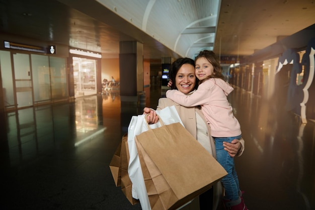 Glückliche Mutter, die Tochter in den Armen hält, sie umarmt, lächelt, in die Kamera blickt und Einkaufstaschen zeigt, die im zollfreien Bereich des Abflugterminals des internationalen Flughafens stehen und auf den Flug warten