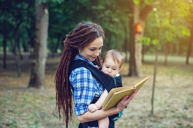 Glückliche Mutter mit einem Baby in einem Riemen ein Buch im Park lesend.