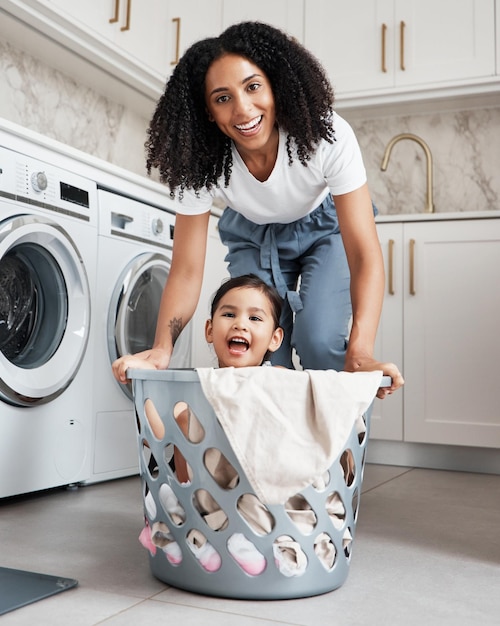 Glückliche Mutter mit ihrem Kind in einem Waschkorb bei sich zu Hause, während sie gemeinsam Wäsche waschen Fröhliche Hausarbeit und Porträt einer jungen Frau, die Spaß mit ihrem Mädchen hat, während sie das Haus putzt