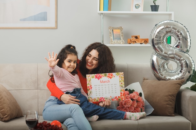 Glückliche Mutter mit ihrer kleinen Tochter, die auf einer Couch mit Blumenstrauß und Kalender des Monats März sitzt und fröhlich im hellen Wohnzimmer lächelt, um den internationalen Frauentag am 8. März zu feiern