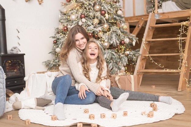 Glückliche mutter mit tochter, die neben dem neujahrsbaum sitzt und den weihnachtsabend zu hause genießt