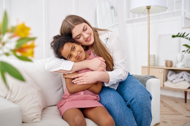 Glückliche Mutter umarmt ihre kleine Tochter auf dem Sofa im Wohnzimmer. Mutter und weibliches Kind verbringen zusammen in ihrem Haus, gute Beziehung, elterliche Fürsorge