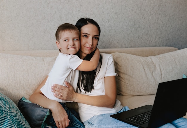 Glückliche Mutter umarmt ihren kleinen Sohn, während sie auf der Couch vor ihrem Laptop sitzt