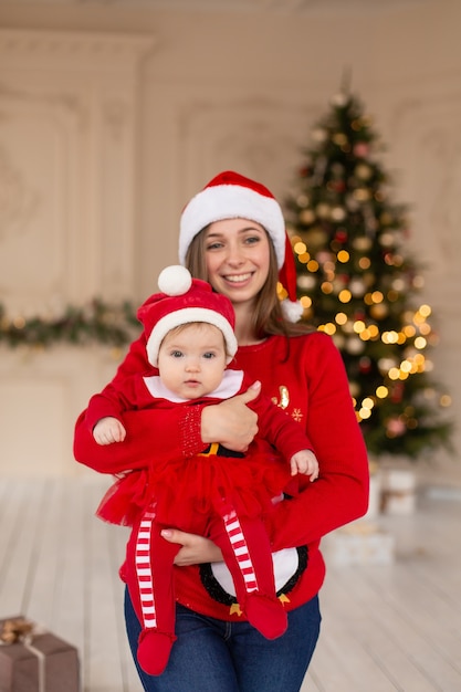 Glückliche Mutter umarmt kleine Tochter im roten Weihnachtsmannkostüm und -hut nahe Weihnachtsbaum.