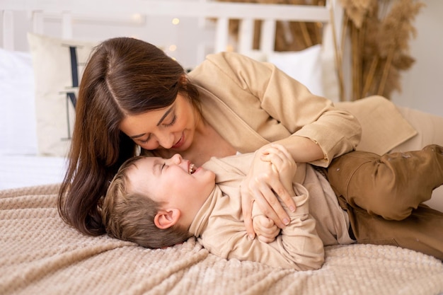 Glückliche Mutter umarmt und küsst ein kleines Kind auf dem Bett in beige-braunen Naturtönen und hat Spaß