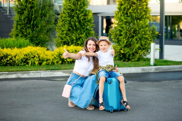 Glückliche Mutter und Baby sitzen auf einem blauen Koffer vor dem Flughafen und fahren in den Urlaub oder reisen zum Flughafen und zeigen der Klasse einen Daumen nach oben