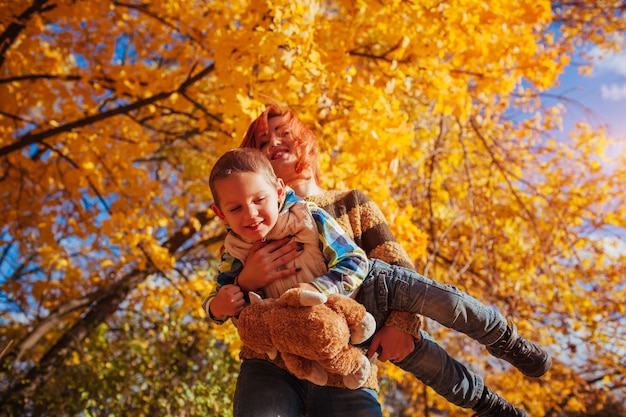 Glückliche Mutter und ihr kleiner Sohn, die im Herbstwald spazieren gehen und Spaß haben. Frau reitet ihr Kind