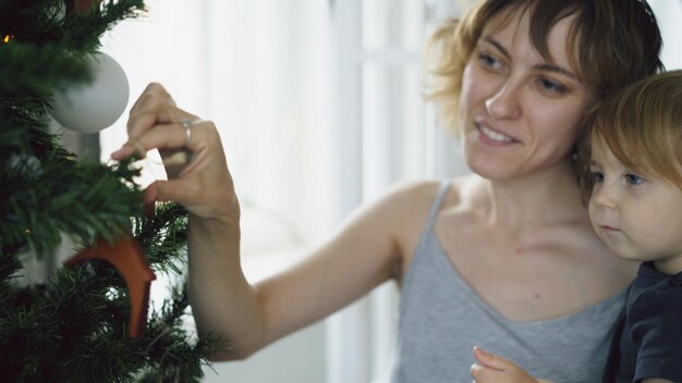 Glückliche Mutter und ihre kleine Tochter spielen zu Hause am Weihnachtsbaum