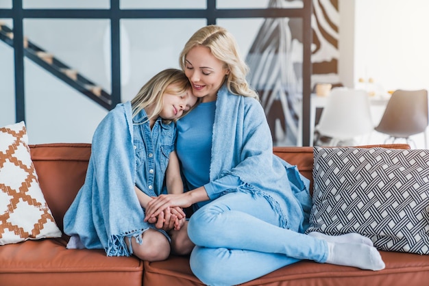 Glückliche Mutter und ihre kleine Tochter umarmen sich zu Hause unter der Decke auf dem Sofa