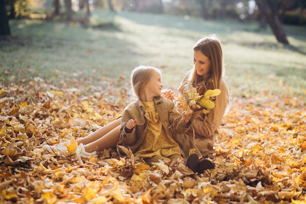 Glückliche Mutter und ihre schöne Tochter sitzen und haben Spaß zwischen den gelben Blättern im Herbstpark.