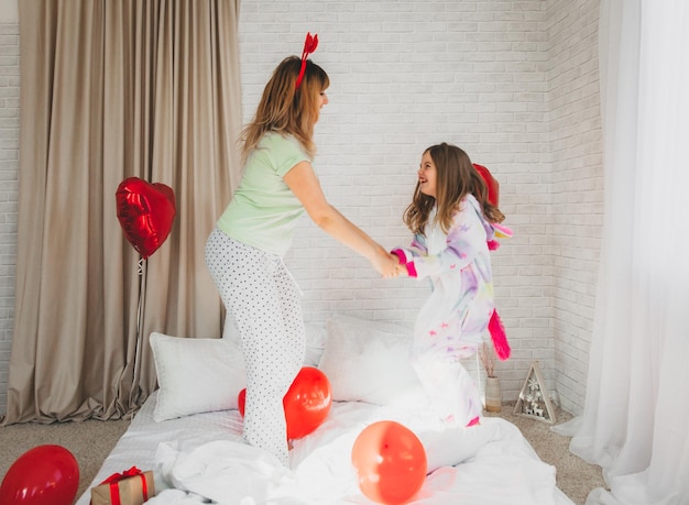 Glückliche mutter und ihre tochter springen auf das bett im schlafzimmer, das für den valentinstag dekoriert ist.