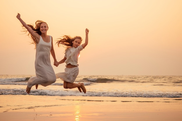 Glückliche Mutter und junge Tochter am Strand bei Sonnenuntergang, die sich am Meer amüsieren und springende und sich freuende Frau und ihr Kind kopieren