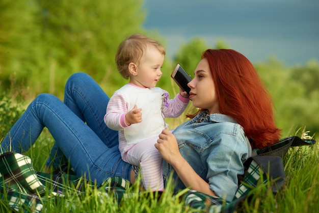 Glückliche Mutter und kleine Tochter, die auf grünem Gras liegen, halten das Handy lächelnd und sehen glücklich und hübsch aus, sitzend auf der Mutter