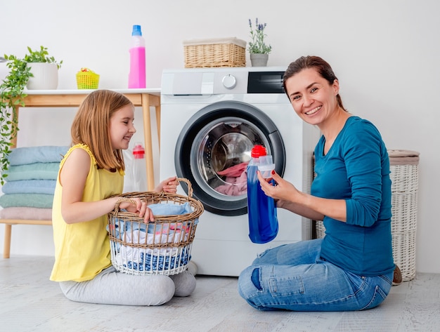 Glückliche Mutter und kleine Tochter, die Wäsche mit Maschine im hellen Raum waschen