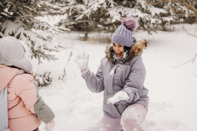 Glückliche Mutter und kleine Tochter spielen Schneebälle in einem verschneiten Naturpark bei Schneefall. Toller lustiger Moment. Gemeinsam Zeit verbringen an einem schönen kalten Wintertag.