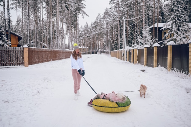 Glückliche Mutter und kleines süßes Mädchen in rosa warmer Oberbekleidung, die Spaß haben reitet aufblasbare Schneeröhre mit rotem Shiba Inu Hund in schneeweißer Winterlandschaft im Freien