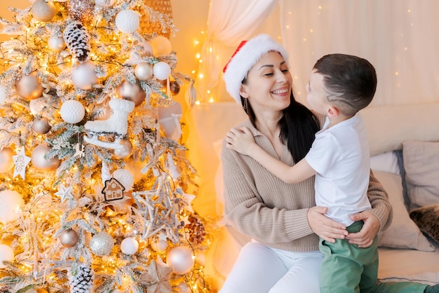 Glückliche Mutter und Sohn tragen Weihnachtsmann Hut kuscheln auf dem Bett im geschmückten Schlafzimmer am Heiligabend in