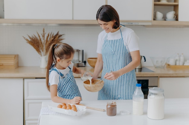 Glückliche mutter und tochter backen zusammen in der küche. verwenden sie verschiedene zutaten. tragen sie schürzen