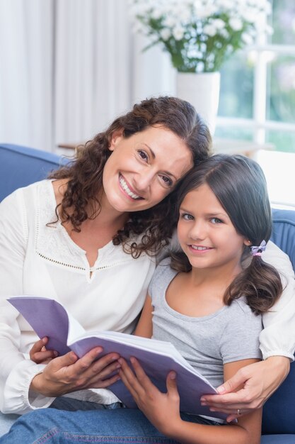 Glückliche Mutter und Tochter, die auf dem Couch- und Lesebuch sitzt