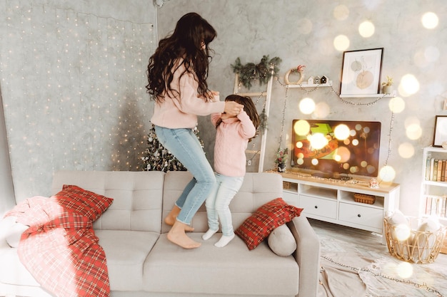 Glückliche Mutter und Tochter, die zusammen auf dem Sofa springen und Händchen halten, Mutter spielt mit niedlichen kleinen Mädchen zu Hause, junge Mutter und Baby lieben es, Zeit miteinander zu verbringen.