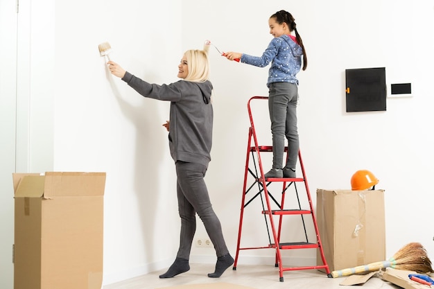 Glückliche Mutter und Tochter machen Reparaturen zu Hause. Lächelnde Frau und Mädchen malen an der Wand im Zimmer.