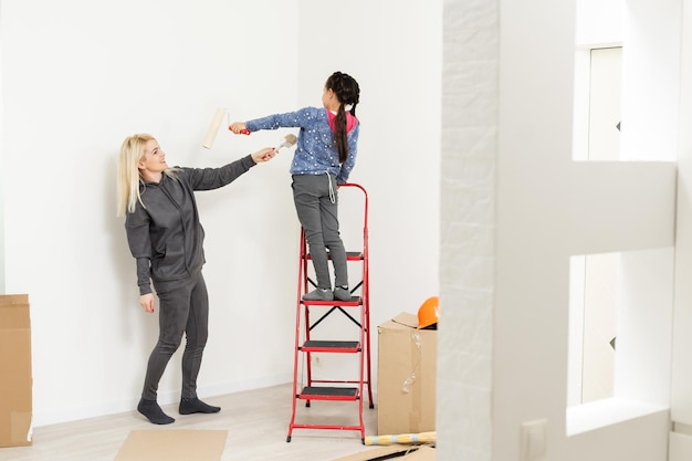 Glückliche Mutter und Tochter machen Reparaturen zu Hause. Lächelnde Frau und Mädchen malen an der Wand im Zimmer.