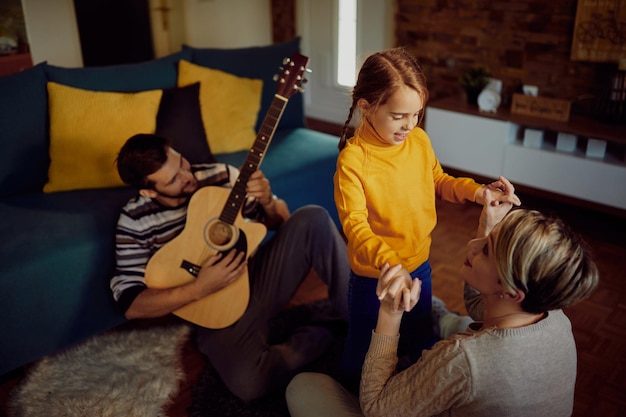 Glückliche Mutter und Tochter unterhalten sich, während Vater zu Hause Akustikgitarre spielt