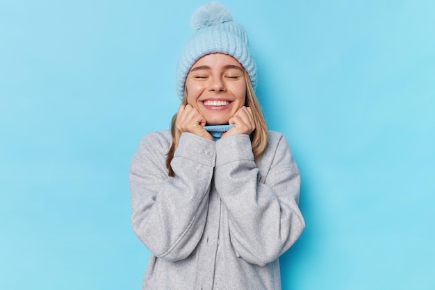 Glückliche optimistische junge Frau schließt die Augen vor Freude lächelt breit zeigt weiße Zähne trägt Strickmütze und Jacke erinnert an angenehme Erinnerungen, die über blauem Hintergrund isoliert sind Menschen und Gefühle