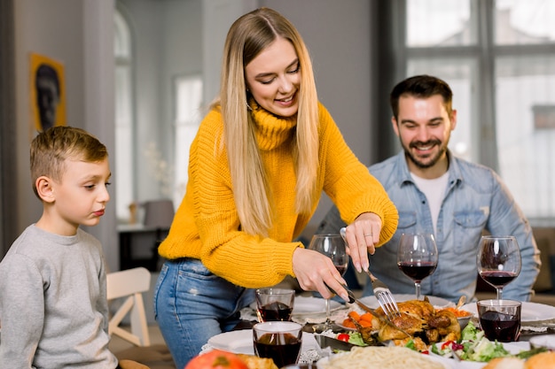 glückliche paar Eltern und ihr kleiner Sohn sitzen am festlichen Tisch und gehen gebratenen Truthahn essen. Glückliche Familie, die köstliches Abendessen zusammen zu Hause hat, während Mutter Truthahn schneidet