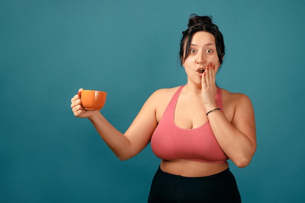 Glückliche plus size positive Frau. Glückliches körperpositives Konzept. Ich liebe meinen Körper. Attraktive übergewichtige Frau posiert vor der Kamera im Studio mit Teetasse in ihren Händen. Mädchen trägt einen Korallen-BH