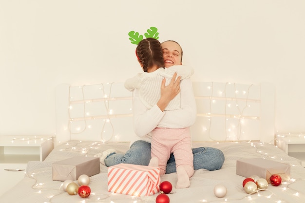 Glückliche positive Mutter und Tochter, die weiße Pullover im lässigen Stil tragen, die mit Weihnachtsdekoration auf dem Bett sitzen und sich zu den Winterferien gratulieren.