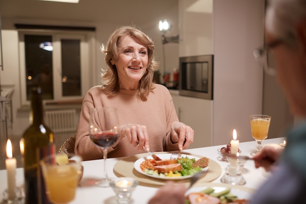 Glückliche reife Frau, die am Tisch sitzt, Abendessen isst und zu ihrem Ehemann während ihrer Verabredung lächelt