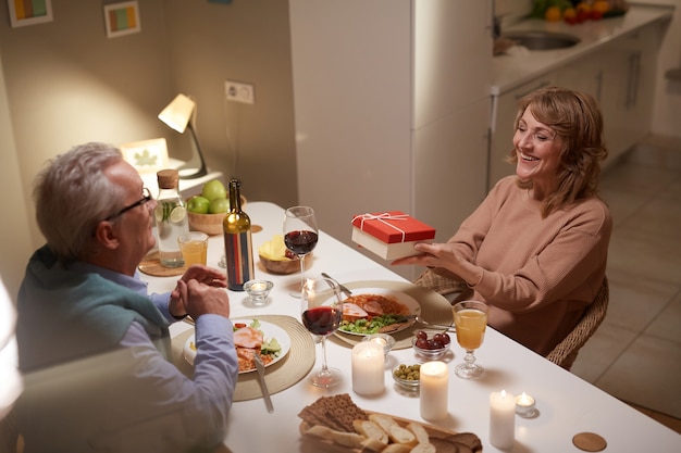 Glückliche reife Frau, die ihrem Ehemann eine Geschenkbox gibt und ihm während des Abendessens zu Hause gratuliert