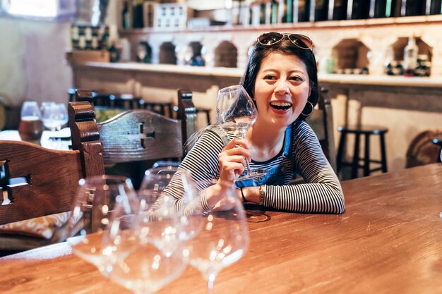 glückliche Reisende, die im Weinkeller im Napa Valley trinkt. Sommelier des jungen Mädchens, der Alkohol in der Feierparty verkostet, die am Holztisch in einem historischen Gebäude sitzt. Frau, die Weinglas hält, lacht