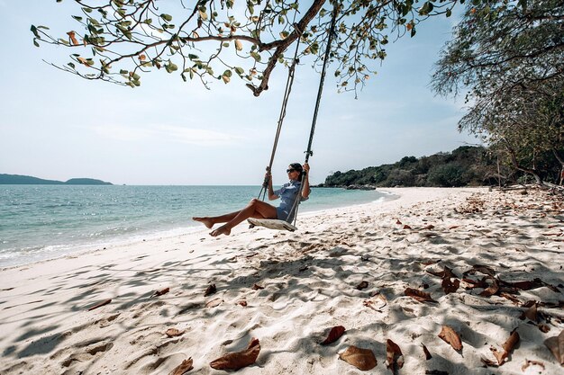 Glückliche reisende Frau in weißem und schwarzem kariertem Kleid und schwarzer Sonnenbrille, die sich auf der Schaukel entspannt. Touristischer Seestrand Thailand, Asien, Sommerferienferienreisereise. Phuket. Thailand