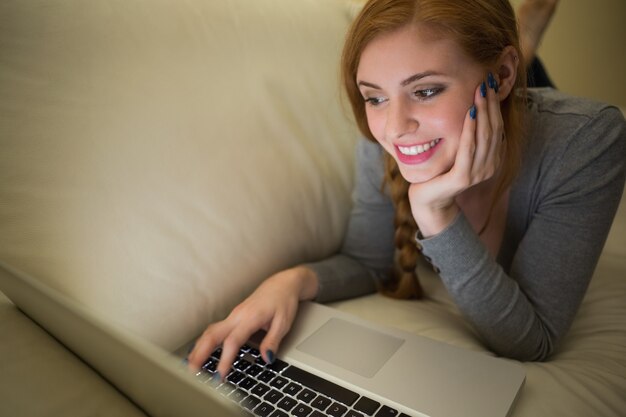 Foto glückliche rothaarige, die auf dem sofa mit ihrem laptop nachts liegt