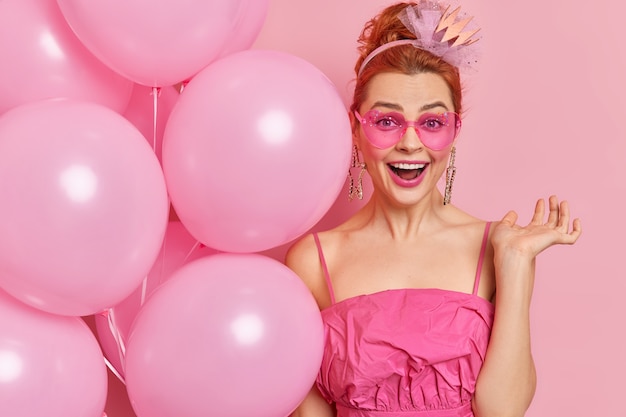Glückliche rothaarige junge Frau trägt trendige herzförmige Sonnenbrille und Kleid hält Haufen aufgeblasener Ballons feiert Geburtstag hat positive Stimmung isoliert über rosa Wand. Festkonzept