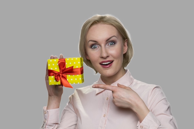 Glückliche schockierte Frau, die auf Geschenkbox zeigt. Aufgeregte Frau, die peresent Box auf grauem Hintergrund hält.