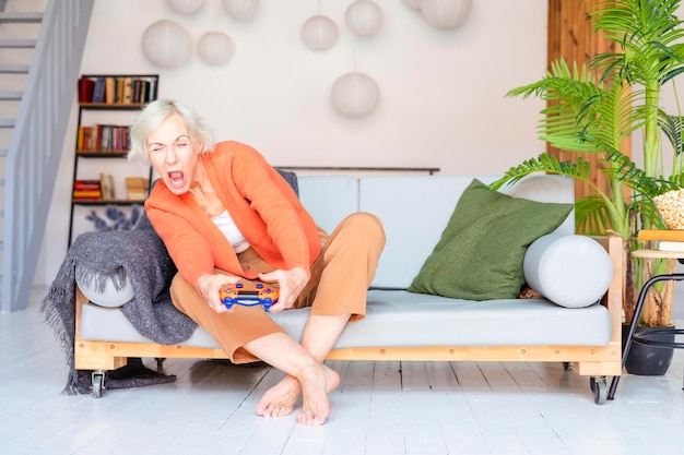 Foto glückliche, schöne, ältere, grauhaarige, ältere frau sitzt auf dem sofa im wohnzimmer, in einer gemütlichen wohnung, lächelnde, ältere frauen mit fernbedienung zum spielen von konsolen, moderne großmutter spielt videospiele online.