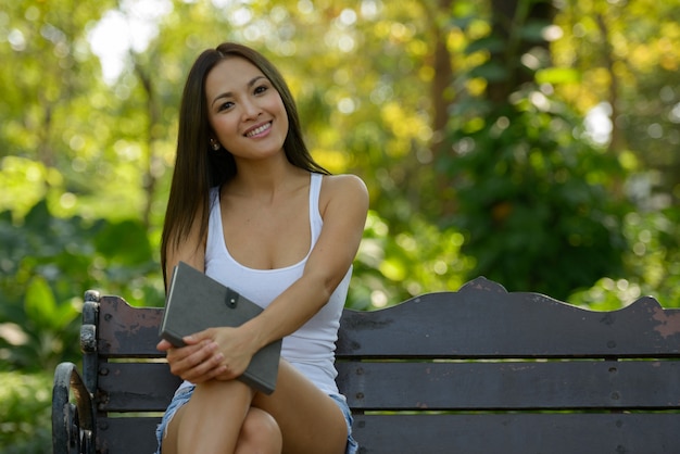 Glückliche schöne asiatische Frau, die Buch am Park im Freien sitzt und hält