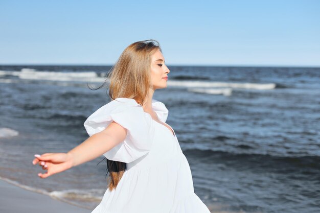 Glückliche, schöne Frau am Meeresstrand, stehend in einem weißen Sommerkleid, offene Arme.