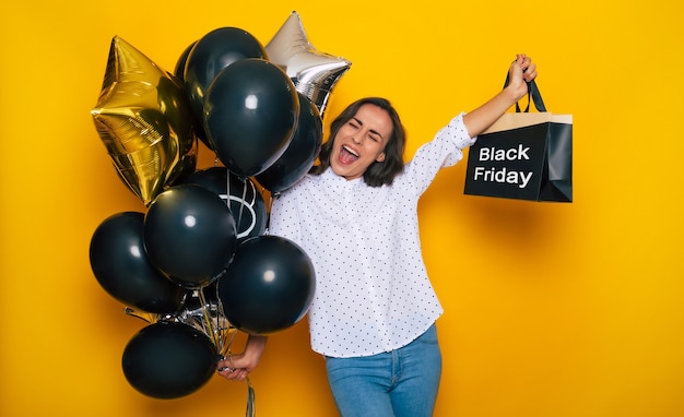 Glückliche schöne Frau, die viele schwarze Luftballons und Einkaufstasche in den Händen hält, während sie am schwarzen Freitag posiert und Kleidung im Einkaufszentrum kauft