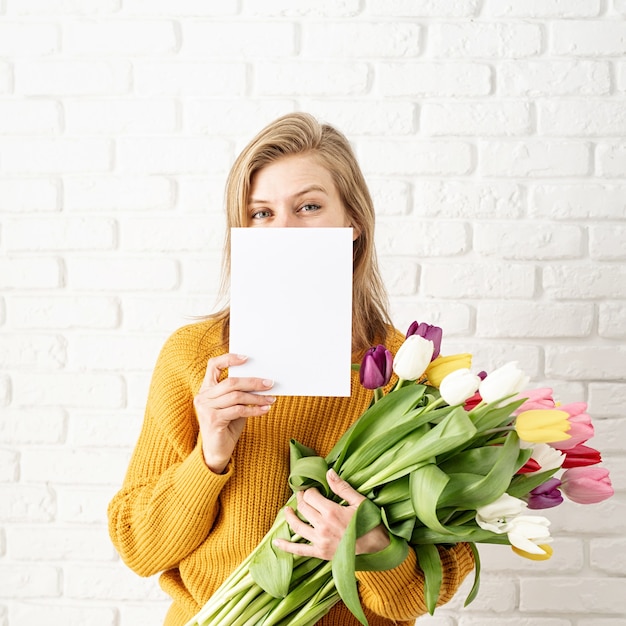 Glückliche schöne Frau in den gelben Kleidern, die Strauß Tulpen und leere Karte halten